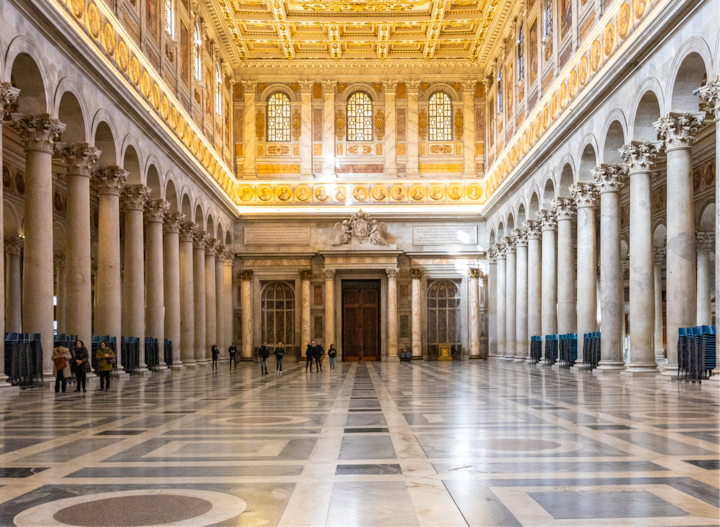 Un espace architectural élégant avec des rangées de grandes colonnes de marbre, des murs décorés, des vitraux et une grande porte en bois au centre.