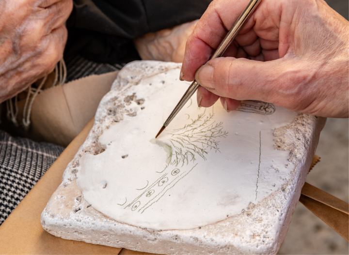 Une personne utilise un pinceau fin pour peindre un arbre sur un carreau de céramique texturé, avec une autre personne assise près d'elle.