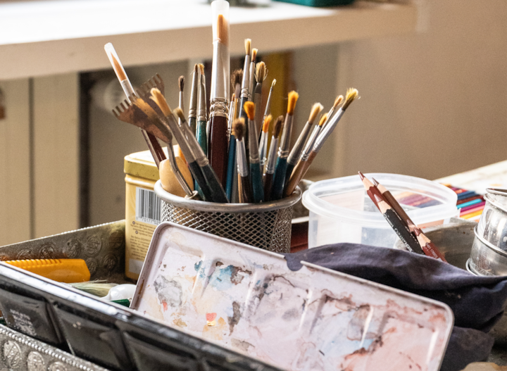 Sur une table de travail, des pinceaux réunis dans un pot en métal ajouré et entouré de fournitures d'artistes telles que des palettes de peinture, des crayons de couleur et des récipients.