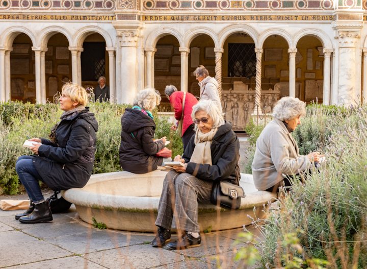 Une cour paisible avec des arches et une architecture décorative en arrière-plan. Plusieurs personnes sont assises sur une fontaine circulaire en pierre entourée de verdure. D'autres personnes marchent non loin de là.