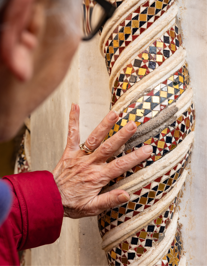 Gros plan sur la main d'une personne âgée touchant une colonne aux motifs en spirale sous forme de mosaïque colorée avec des carreaux rouges, dorés, noirs et blancs, évoquant une expérience tactile.