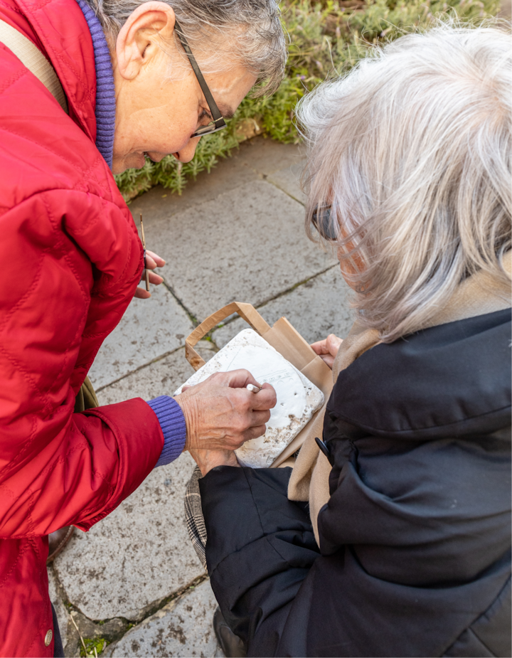 Deux personnes en extérieur, l'une dessine sur un carreau de céramique tandis que l'autre observe. Un sac en papier repose sur ses genoux.
