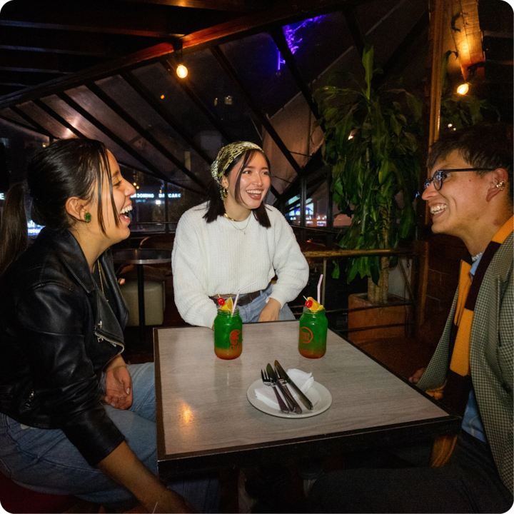 Trois personnes sont assises autour d'une petite table dans un café à l'éclairage tamisé, avec deux cocktails colorés garnis de fruits devant elles. À l'arrière-plan, une plante et de grandes baies vitrées par lesquelles on aperçoit les lumières de la ville.