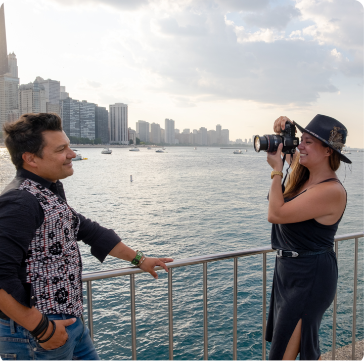 Une personne portant une robe noire et un chapeau prend en photo une autre personne debout au bord de l'eau. Au loin se dessinent des bateaux et la silhouette d'une ville sous un ciel nuageux.