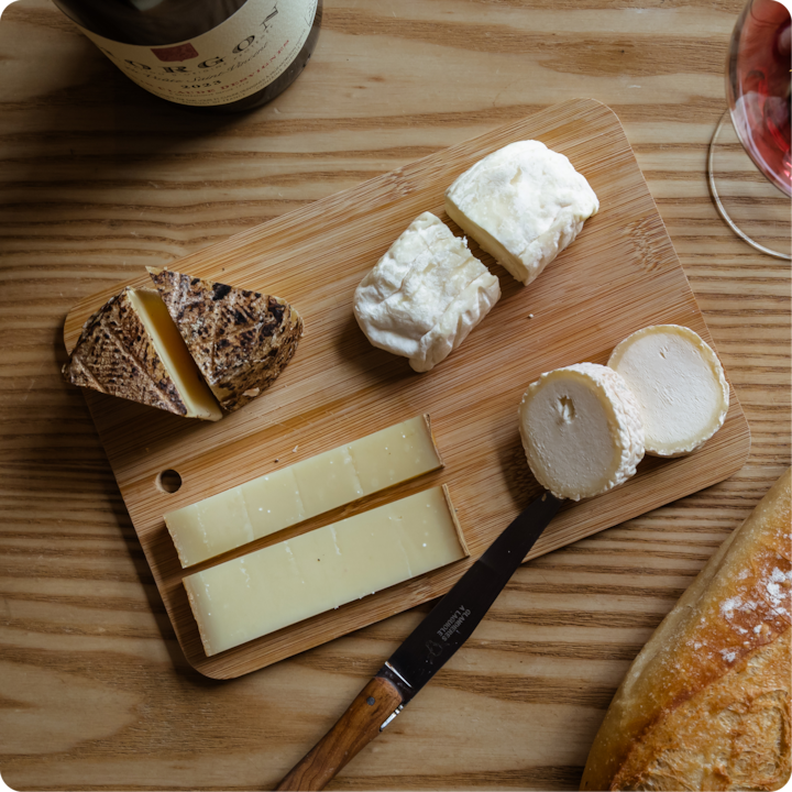 Une planche en bois sur laquelle sont présentés divers fromages à pâte dure, à pâte molle et affinés, entourée d'une bouteille de vin, d'un verre de vin rouge et d'une miche de pain, suggérant une expérience culinaire rustique.