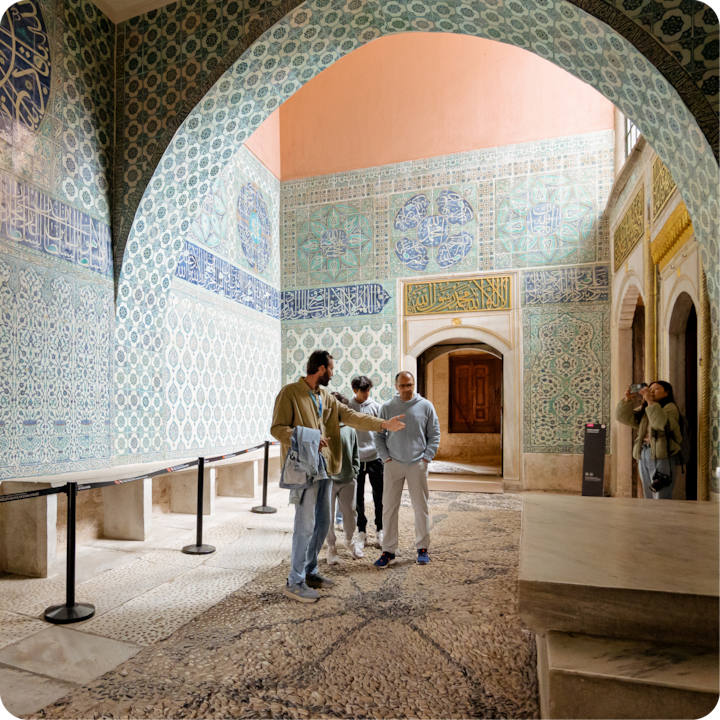 Un groupe de personnes se trouve dans une pièce richement décorée, avec des carreaux aux motifs géométriques complexes bleus et verts, de la calligraphie arabe sur les murs et un haut plafond voûté. Une personne fait des gestes en discutant avec les autres.