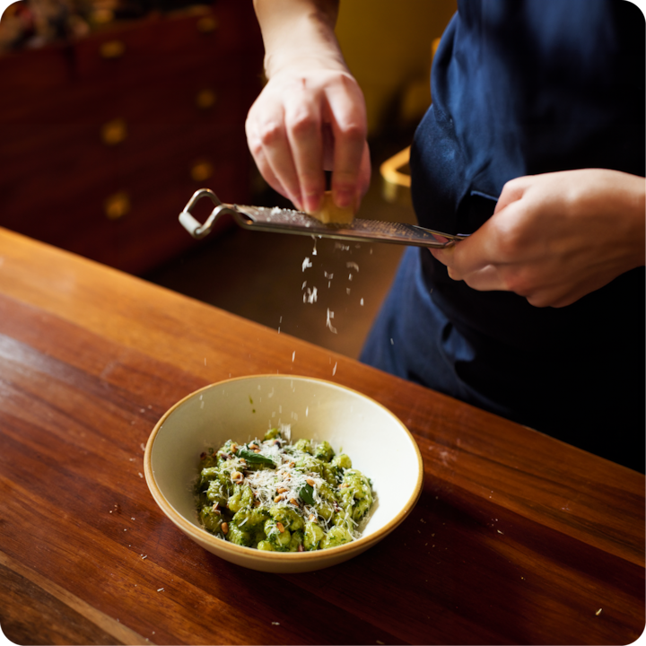 Quelqu'un râpe du fromage frais au-dessus d'un bol de pâtes garnies d'une sauce verte, d'herbes et de noix, le tout posé sur un plan de travail en bois.