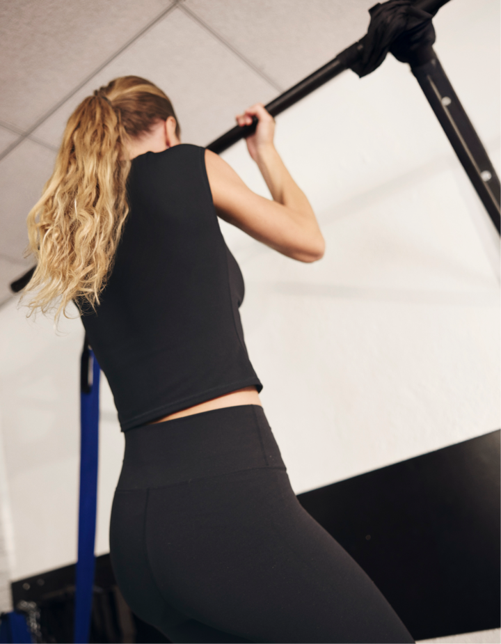 Une femme blonde coiffée d'une queue de cheval et portant des vêtements de sport noirs s'exerce à l'aide d'une barre de traction noire dans un espace intérieur lumineux.