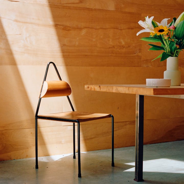 Dans une pièce ensoleillée aux murs en bois, une chaise en bois avec un dossier incurvé et un cadre en métal noir, placée à côté d'une table en bois sur laquelle repose un bouquet de fleurs dans un vase.