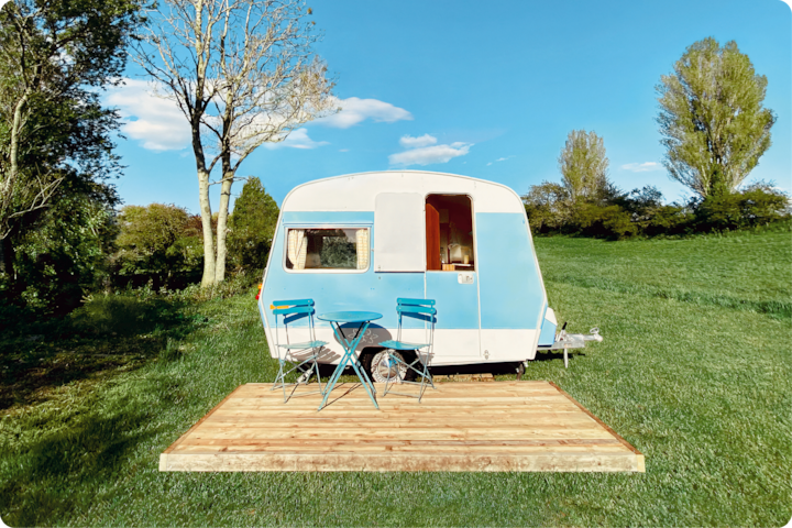 Un petite caravane bleue et blanche est garée dans un champ verdoyant. Devant, une estrade en bois accueille une table turquoise et deux chaises assorties, sous un ciel dégagé.