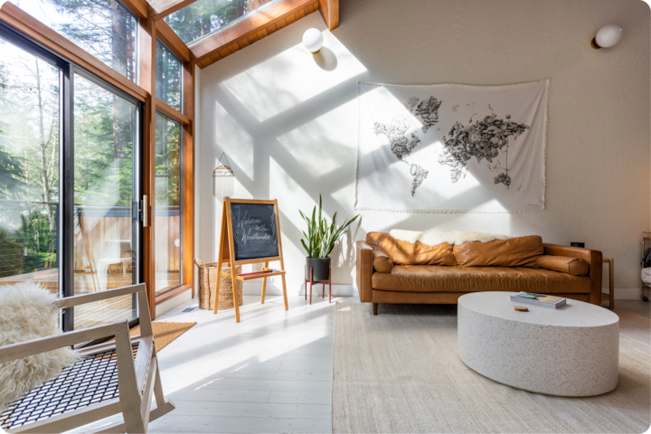 Un salon lumineux et moderne avec de grandes baies vitrées, un canapé en cuir fauve, une table basse ronde et blanche et une tapisserie murale représentant une carte du monde. Un tableau noir de bienvenue aux voyageurs placé à côté d'une plante en pot et une chaise en bois.
