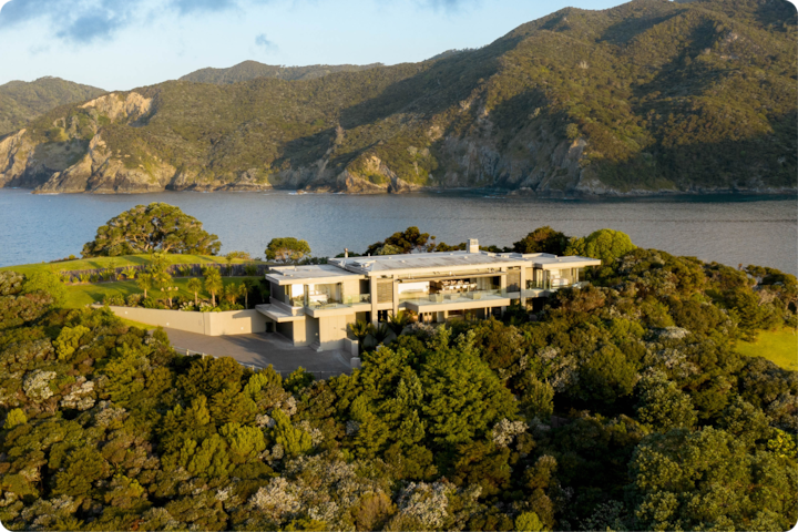 Une maison moderne luxueuse entourée d'une végétation luxuriante sur une colline surplombant un lac paisible avec des montagnes en arrière-plan, au coucher du soleil.