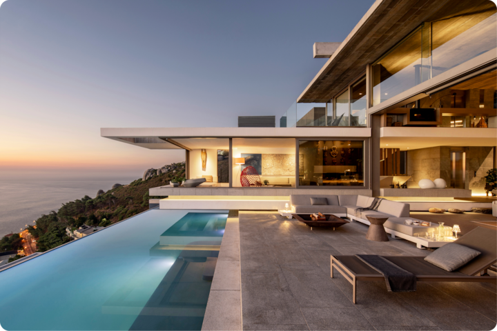 Maison moderne luxueuse au coucher du soleil, avec une piscine à débordement surplombant une colline sur la côte, un salon d'extérieur et des pièces intérieures éclairées.