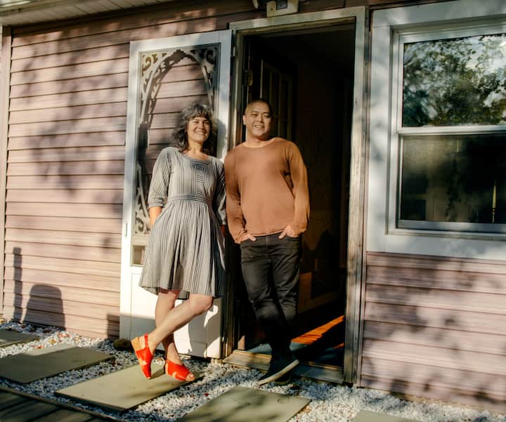 A cozy house entrance with two individuals standing by the open door, one wearing a striped dress and red shoes, and the other in a brown sweater and black pants, with sunlight filtering in.