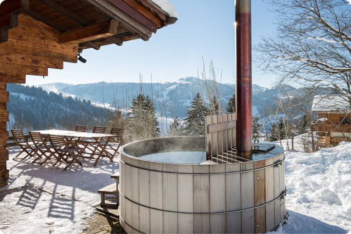 Jacuzzi extérieur en bois dont s'échappe de la vapeur dans un paysage de montagne enneigé surplombant une forêt pittoresque. À proximité du jacuzzi, on aperçoit le toit d'une cabane ainsi qu'une table et des chaises en bois.