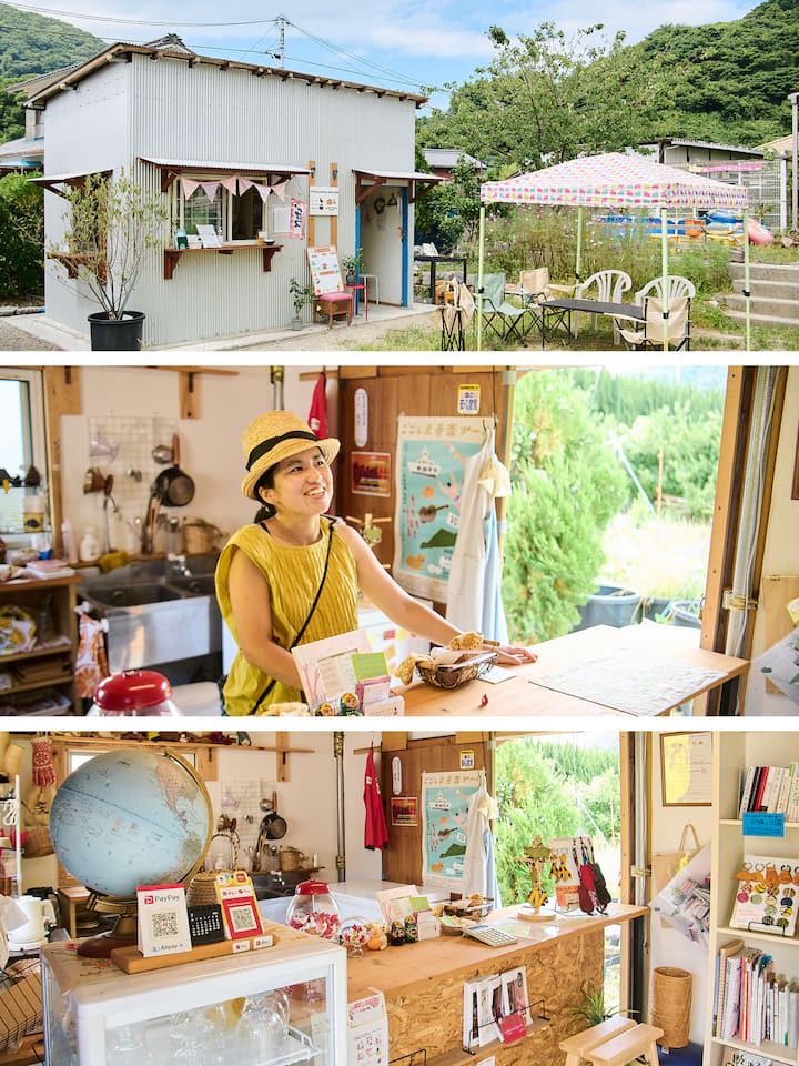 A small countryside cafe with a metal exterior, colorful banners, a seating area under a patterned canopy, and lush greenery surrounding it.