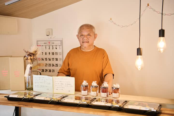 A table displaying neatly arranged prepared meal boxes and jars of preserves, with handwritten notes in Japanese offering details about the items, under warm lighting in a cozy interior space.