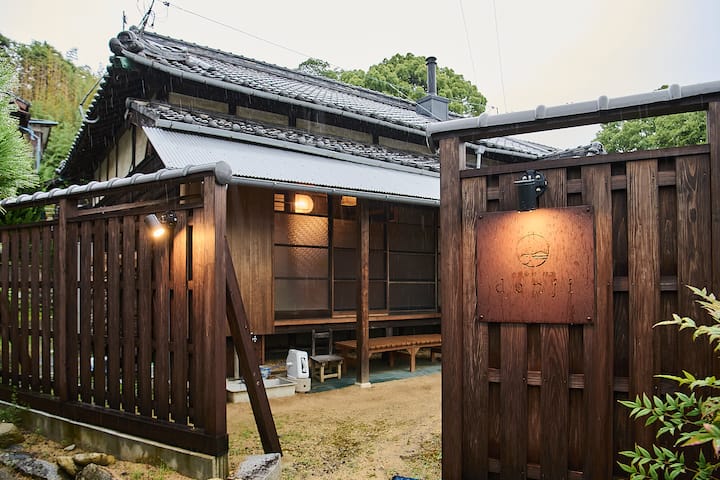 Traditional Japanese wooden house with a tiled roof surrounded by greenery, featuring a wooden sign with carved text near the entrance.