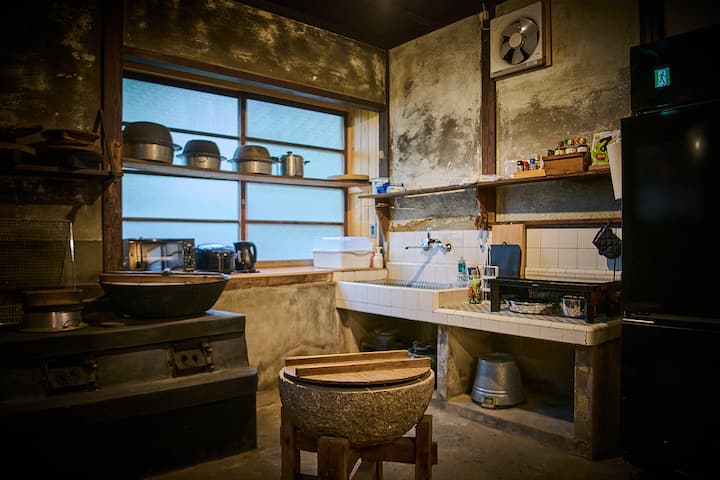 Traditional Japanese kitchen with a rustic stone stove, wooden mortar, and shelves holding pots and utensils near a frosted glass window.