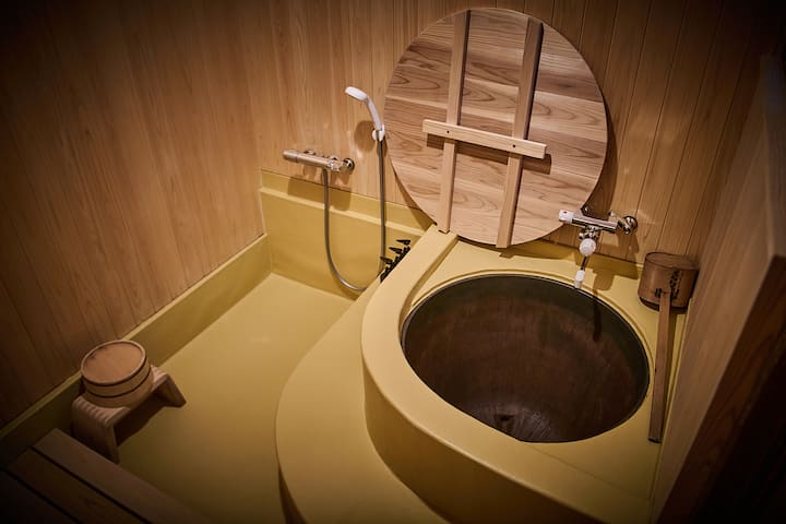 Traditional Japanese wooden bathroom featuring a round soaking tub with a wooden lid, handheld showerhead, and small stool on a yellow floor.