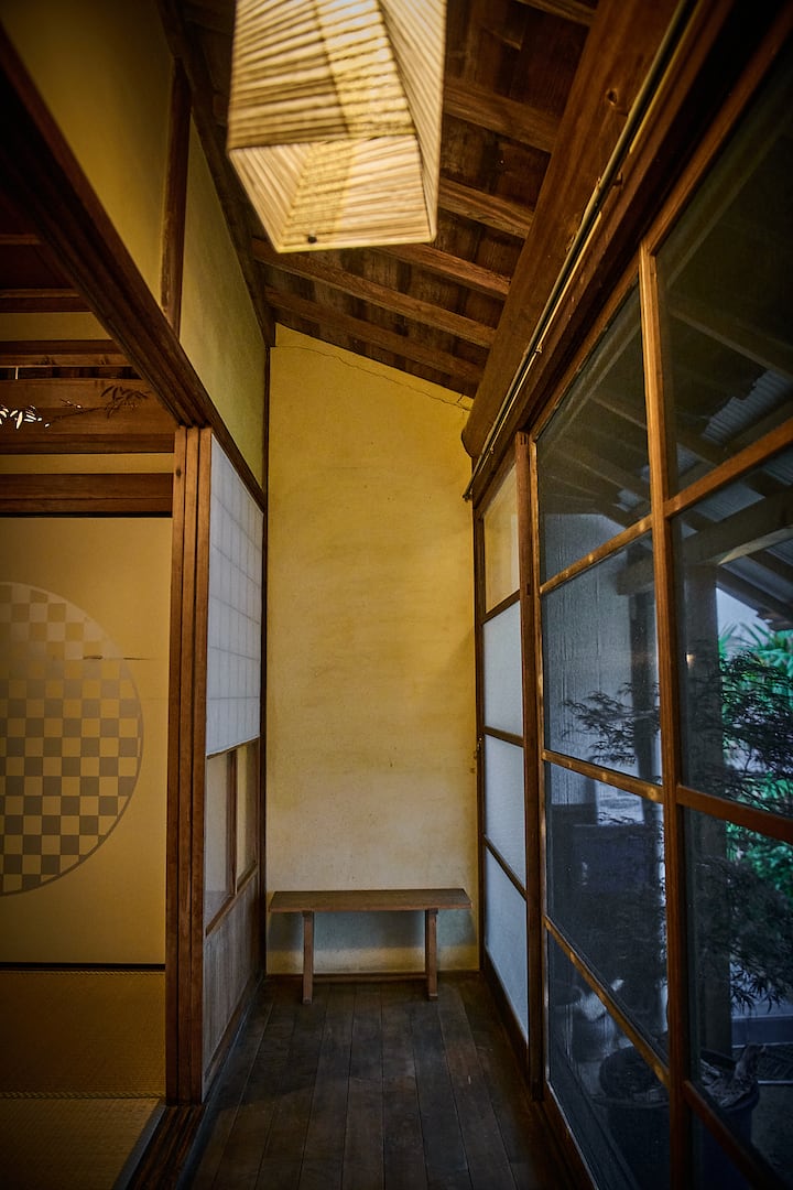 A hallway in a traditional Japanese home with wooden floors and walls, a hanging paper lantern, and glass sliding doors revealing greenery outside.