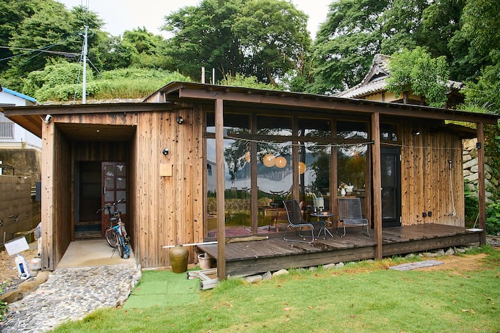 A traditional Japanese wooden house with large glass windows, surrounded by lush greenery. The patio features a small table and chairs, offering a cozy outdoor seating area.