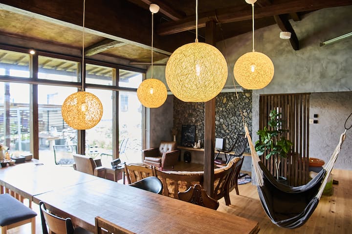 A cozy, modern Japanese living and dining space featuring wooden beams, warm yellow pendant lights, a hammock, and large windows overlooking a garden.