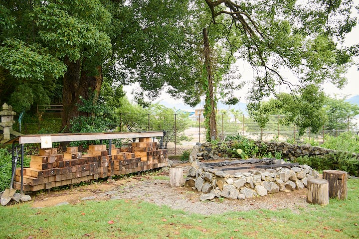 A serene outdoor space in Japan featuring a woodpile under a metal roof and a stone fire pit surrounded by tree stumps used as seating, with lush greenery and trees in the background.
