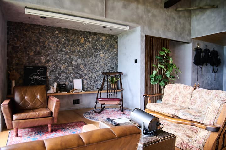 Cozy living room in Japan featuring rustic decor, a stone accent wall, vintage armchairs, a patterned rug, and a rocking chair surrounded by indoor plants.