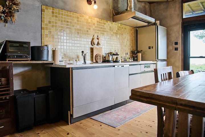 A cozy kitchen in Japan with a modern design, featuring a yellow tiled backsplash, wooden flooring, and a dining table. The window offers a view of greenery outside.