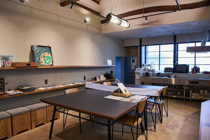 A well-lit community space featuring a modern kitchen and dining area in Japan, with wooden tables, chairs, and shelves displaying local items.