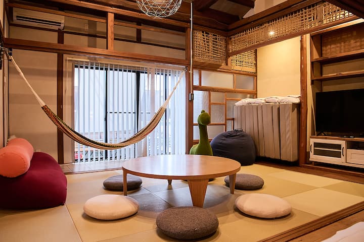A cozy Japanese-style living room with tatami mats, a round wooden table surrounded by cushions, a hammock hanging near a sliding window, and a corner with a TV and storage shelves.