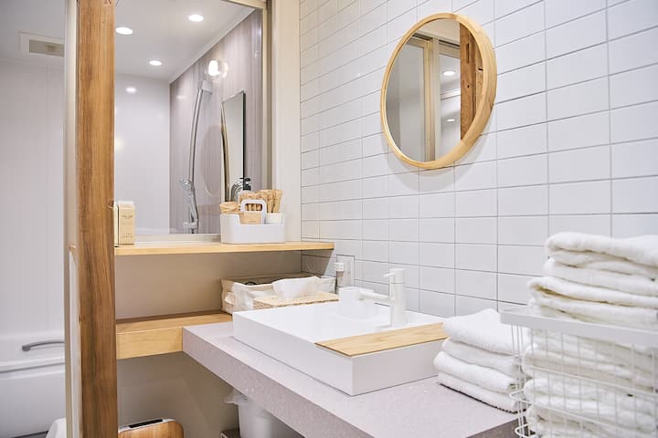 A modern bathroom with a round wooden-framed mirror, a white tiled wall, a countertop with neatly arranged toiletries, a sink with a wooden tray, and folded white towels on a wire rack.