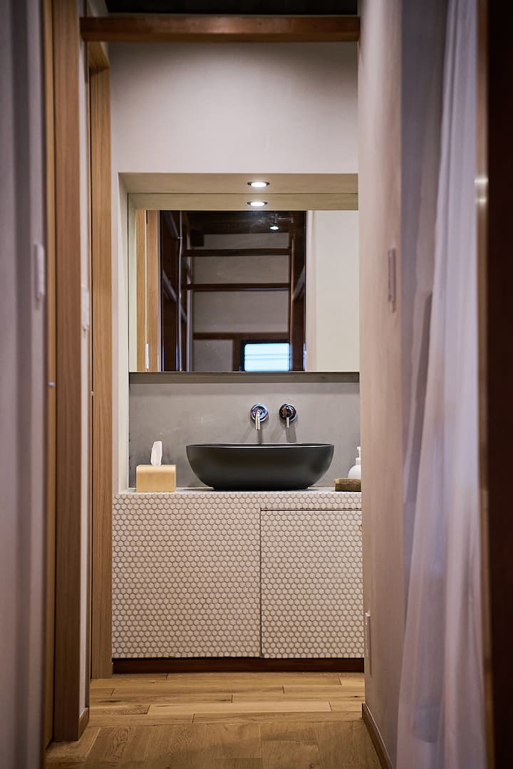 A minimalist Japanese-style bathroom featuring a honeycomb-patterned sink cabinet, a black bowl sink, and a wooden-framed mirror reflecting interior details.