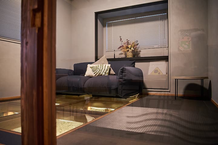 A cozy modern room in Japan with a navy sofa decorated with cushions, a small vase with dried flowers on the windowsill, and a glass floor panel revealing an illuminated display underneath.