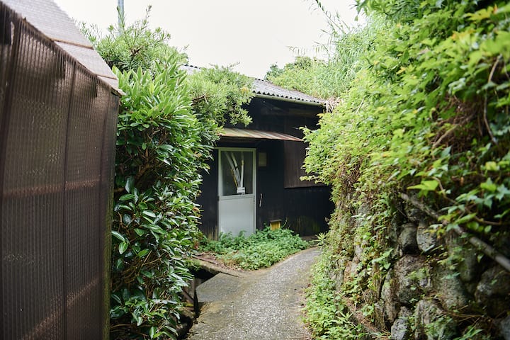 Narrow pathway surrounded by lush greenery leading to a traditional Japanese house with a wooden exterior and a white door.