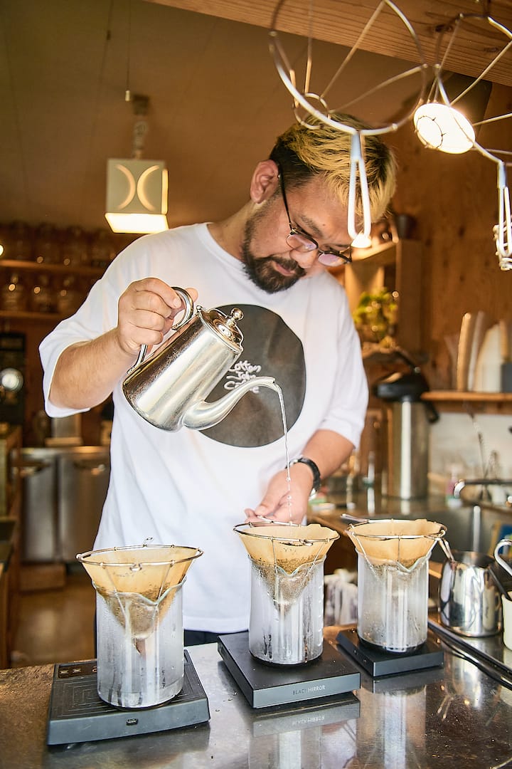A person wearing a white shirt pouring hot water from a silver kettle into a pour-over coffee setup with three glass drippers on digital scales in a cozy, wooden kitchen environment.