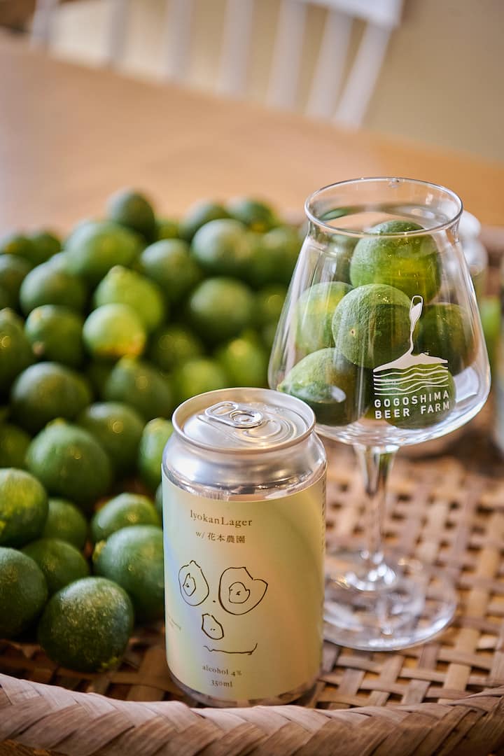 A can of Iyokan Lager beer labeled with an illustration of citrus fruit, placed on a woven tray beside a glass labeled 'Gogoshima Beer Farm' filled with green citrus fruits.