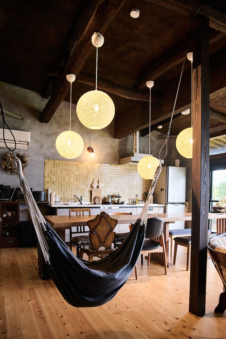 A cozy kitchen and dining area with wooden beams, warm yellow tiled backsplash, hanging spherical lights, dining table with chairs, and a black hammock hanging in the foreground.