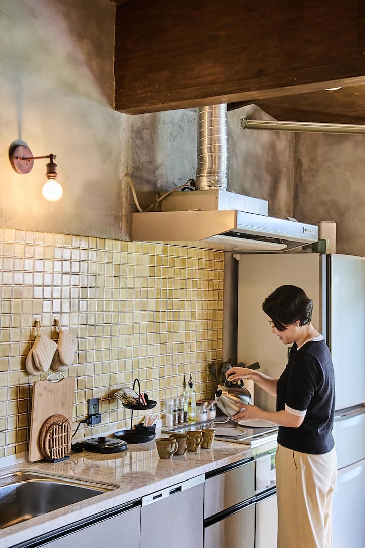 A person standing in a cozy, modern kitchen with yellow tiled backsplash, pouring hot water from a kettle into a cup, with neatly arranged kitchenware on the counter.