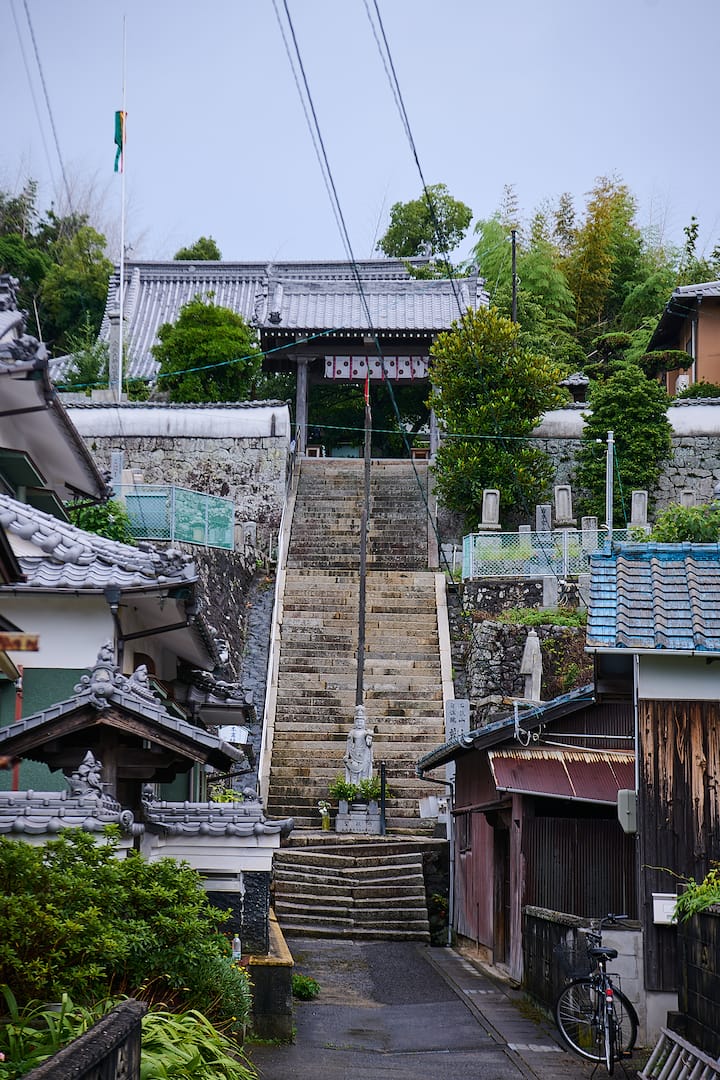 Stone staircase leading to a traditional Japanese temple gate, surrounded by greenery and tiled-roof houses, with a small stone statue at the base of the steps.