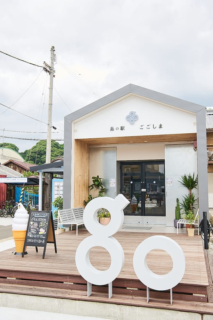 Entrance of a small modern building with a sign reading '島の駅 ごごしま' in Japanese, surrounded by plants, featuring a wooden deck with large white '8' and '0' sculptures in front.