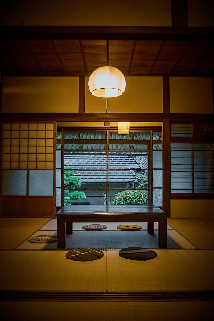 A traditional Japanese tatami room with a low wooden table, round floor cushions, paper sliding doors, and a view of a garden with a tiled roof.