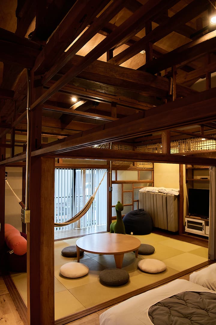 Traditional Japanese-style living space with tatami flooring, a round wooden table surrounded by cushions, a hammock by the window, and a loft area with wooden beams and a cozy ambiance.