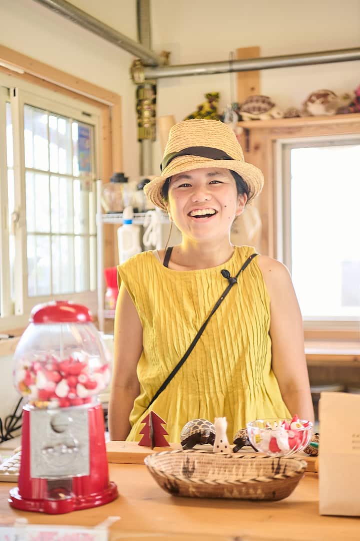 A person wearing a yellow sleeveless dress and a straw hat standing behind a wooden counter with small decorative items, including a red gumball machine, wooden figurines, and a woven basket, in a well-lit space with large windows.