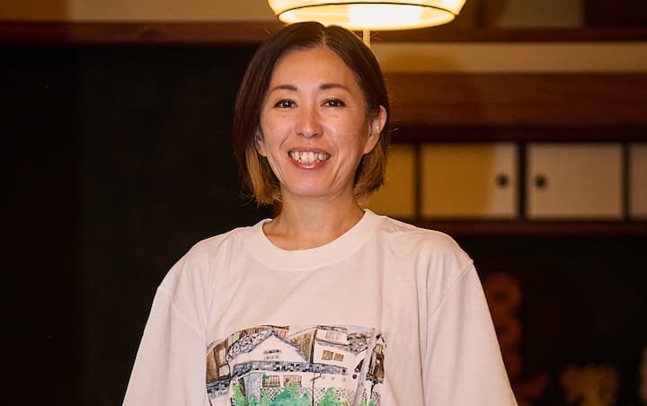 A person wearing a white T-shirt with an illustrated design of a traditional Japanese house, standing inside a warmly lit room with wooden beams and a traditional aesthetic.
