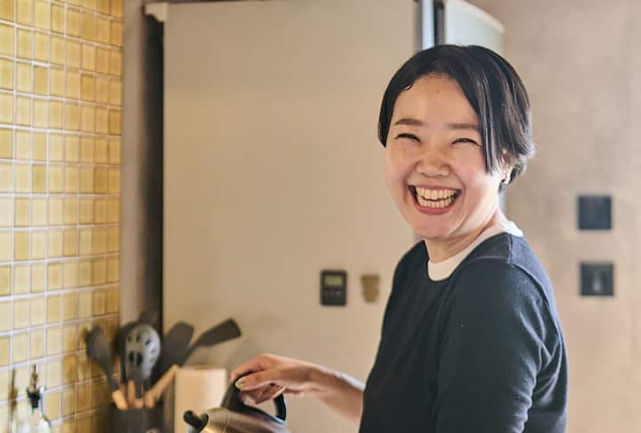 A person standing in a cozy kitchen pouring water from a kettle, with tiled walls and cooking utensils visible in the background.