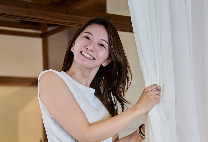 A woman in a sleeveless white top standing inside a traditional-style guest house, holding a white curtain open, revealing a wooden interior frame.