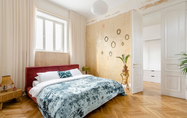 Une chambre lumineuse et élégante avec un cadre de lit en velours rouge, des draps blancs et une couette à motifs bleus. La pièce est égayée par un mur beige texturé avec des décorations rondes, de grandes fenêtres encadrées de rideaux blancs et du parquet. Une porte mène à une pièce attenante avec des rangements.