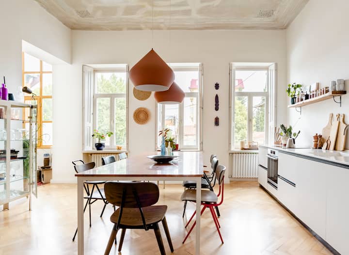 Cuisine lumineuse et moderne avec de grandes fenêtres, une table en bois entourée de chaises dépareillées et des suspensions en terre cuite.